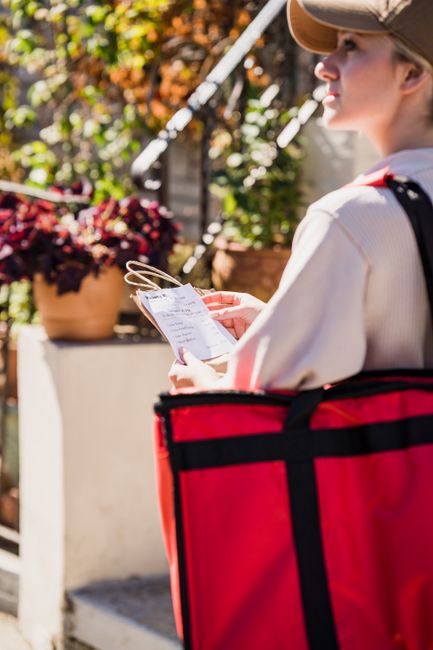 A woman dressed in a light-colored jacket and beige cap, holding a printed flower delivery order or receipt, standing outdoors on a sunny day. She is next to a vibrant pink and red thermal delivery ba