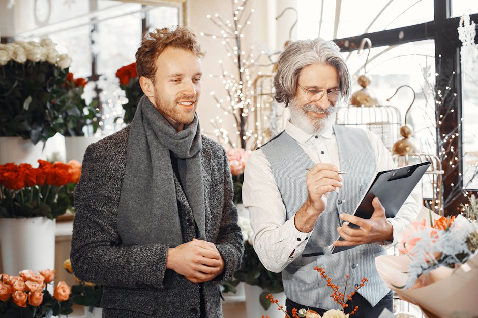 In a bright, well-lit flower shop, a young man with light brown hair and a beard, dressed in a dark grey coat and matching scarf, stands next to an older man with grey hair, glasses, and a grey waistc