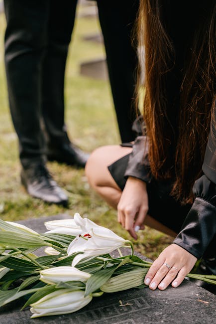 A person in a black outfit is kneeling on a grave, placing a bouquet of white lilies with long green leaves on the headstone. The lilies have delicate petals with a smooth texture and are arranged in