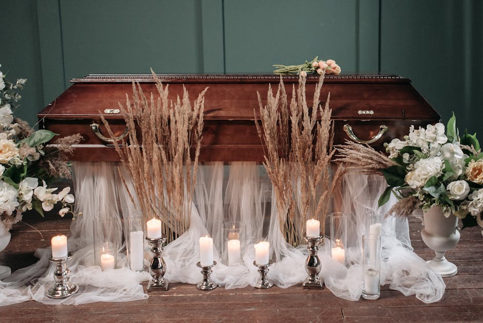 A wooden funeral casket surrounded by floral arrangements and candles, including white roses, orchids, and peach-colored flowers, with beige pampas grass placed in front of the casket. The floral disp