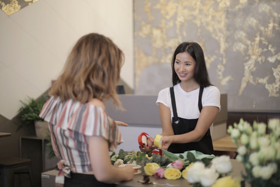 A smiling floral shop assistant with long dark hair, wearing a white t-shirt and black apron, is handing a small roll of red ribbon to a female customer with shoulder-length blonde hair dressed in a s